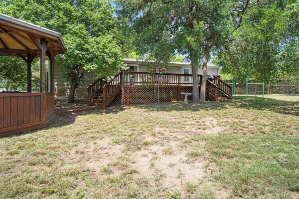 A wooden deck with a staircase leading to a covered area, surrounded by trees and a fenced yard. - Kingfisher | 3BR Tarpley Cabin with Views & Trails (Tarpley)