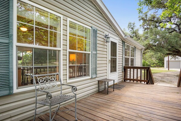 A wooden deck with a bench and a table, a house with large windows and shutters, and a tree in the background.