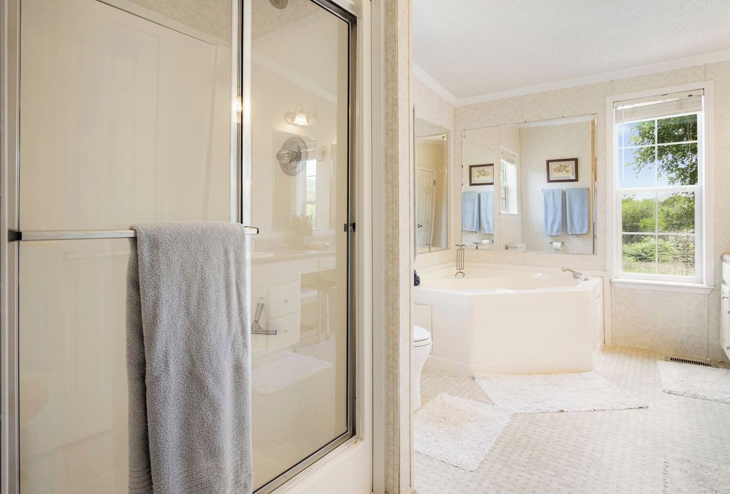 A large en suite bathroom featuring a walk-in shower with glass doors and a separate corner soaking tub, accented by large mirrors and a window overlooking the property.