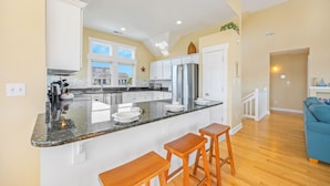 A modern kitchen with white cabinets, a stainless steel refrigerator, and a large island with bar stools. - Oceanview Home w/ Private Pool, Hot Tub, Game Room, Theater, and Volleyball (Waves)