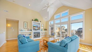 A spacious living room with two blue sofas, a fireplace, and a large window offering a view of the neighborhood. - Oceanview Home w/ Private Pool, Hot Tub, Game Room, Theater, and Volleyball (Waves)