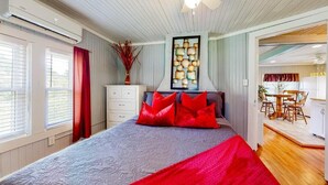 Una camera da letto con un letto, una ventola a soffitto, una finestra con persiane, una tenda rossa, un vaso con piante essiccate e un'area pranzo con tavolo e sedie.