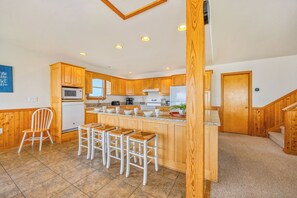 A modern kitchen with wooden cabinets, a central island with bar stools, and a microwave above the sink. - Soundfront Home in Rodanthe w/ Hot Tub, Convenient Windsports Access, Game Room (Rodanthe)