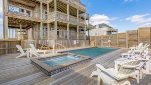 Een houten terras met een jacuzzi en lounge stoelen, een houten omheining en een gebouw met meerdere balkons.