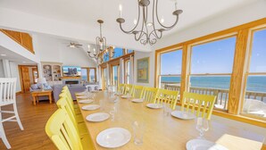 Een eetgedeelte met een lange tafel voor zes personen, gele stoelen en uitzicht op de zee door grote ramen.