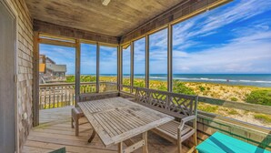 Une terrasse en bois avec une table et des chaises, donnant sur une plage bordée de dunes et où l’on peut admirer les vagues de l’océan.