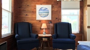 A cozy room with wooden walls, two blue armchairs, a small table with a lamp, and a framed picture of a beach scene. - Diane Cottage, York Manor, Short Sands Beach (York)
