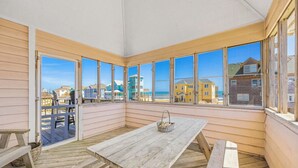 A covered patio with wooden benches and a picnic table, large windows offering a view of colorful houses and the beach. - Semi-Oceanfront Dog Friendly Home w/ Private Pool, Hot Tub, Foosball, Basketball (Rodanthe)
