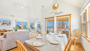 A dining area with a table set for six, a sofa, and a view of the sea through large windows. - Semi-Oceanfront Dog Friendly Home w/ Private Pool, Hot Tub, Foosball, Basketball (Rodanthe)