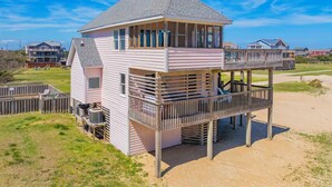 A two-story beach house with a balcony, a small deck, and a visible air conditioning unit. - Semi-Oceanfront Dog Friendly Home w/ Private Pool, Hot Tub, Foosball, Basketball (Rodanthe)