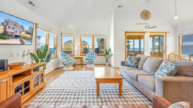 A spacious living room with a large window, a ceiling fan, a wooden coffee table, and a sofa with patterned cushions. - Semi-Oceanfront Dog Friendly Home w/ Private Pool, Hot Tub, Foosball, Basketball (Rodanthe)