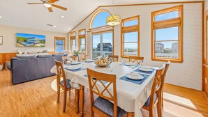 Comedor con mesa dispuesta para seis, sillas de madera, ventilador de techo y ventanas amplias con vista a casas.