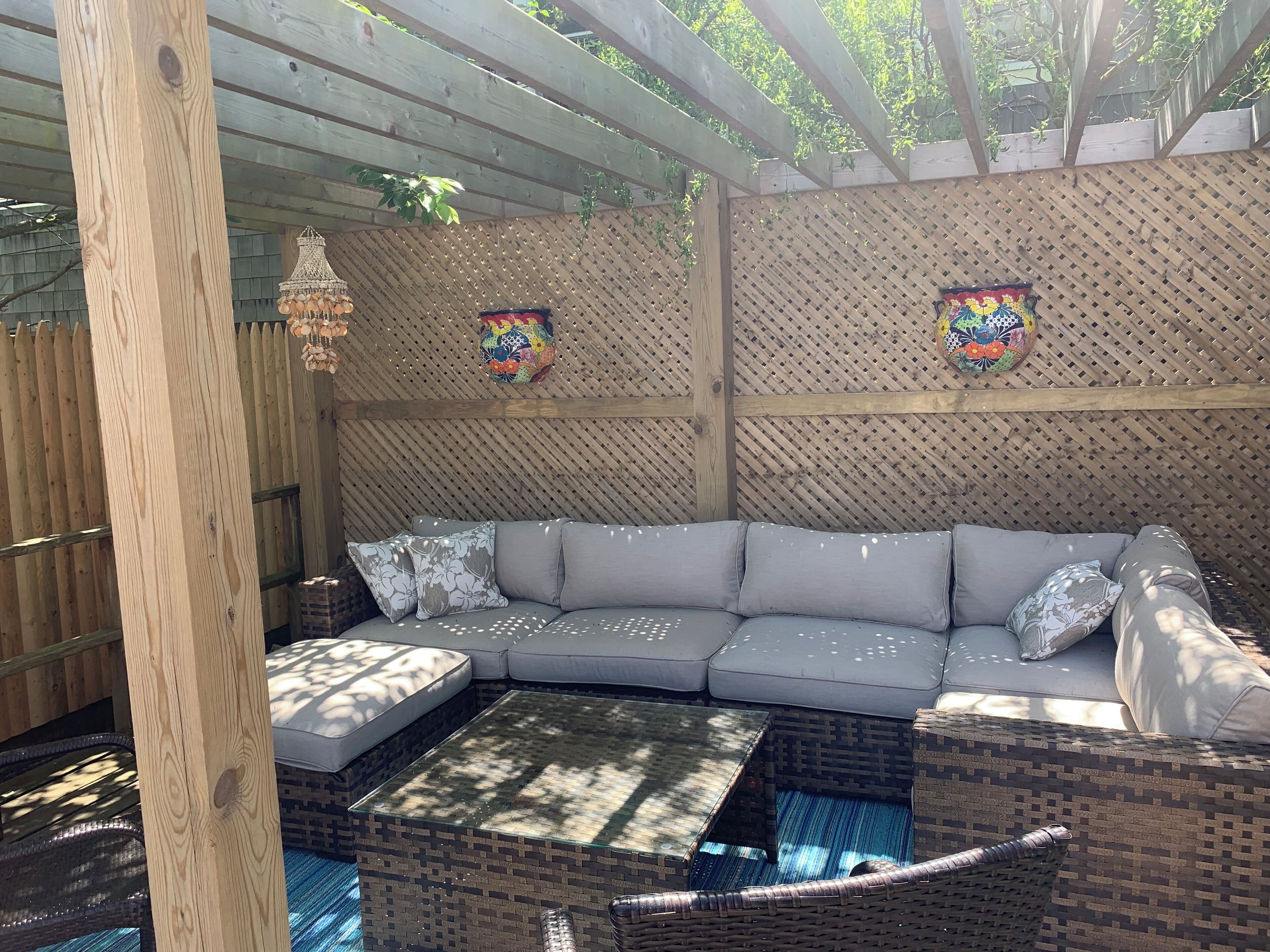 A covered outdoor seating area with a cushioned sofa, a glass-top coffee table, and a wicker side table.