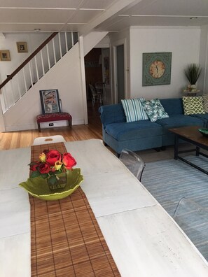 Una sala de estar con un sofá azul, una mesa de centro, una mesa de comedor con un centro de mesa floral y una escalera.