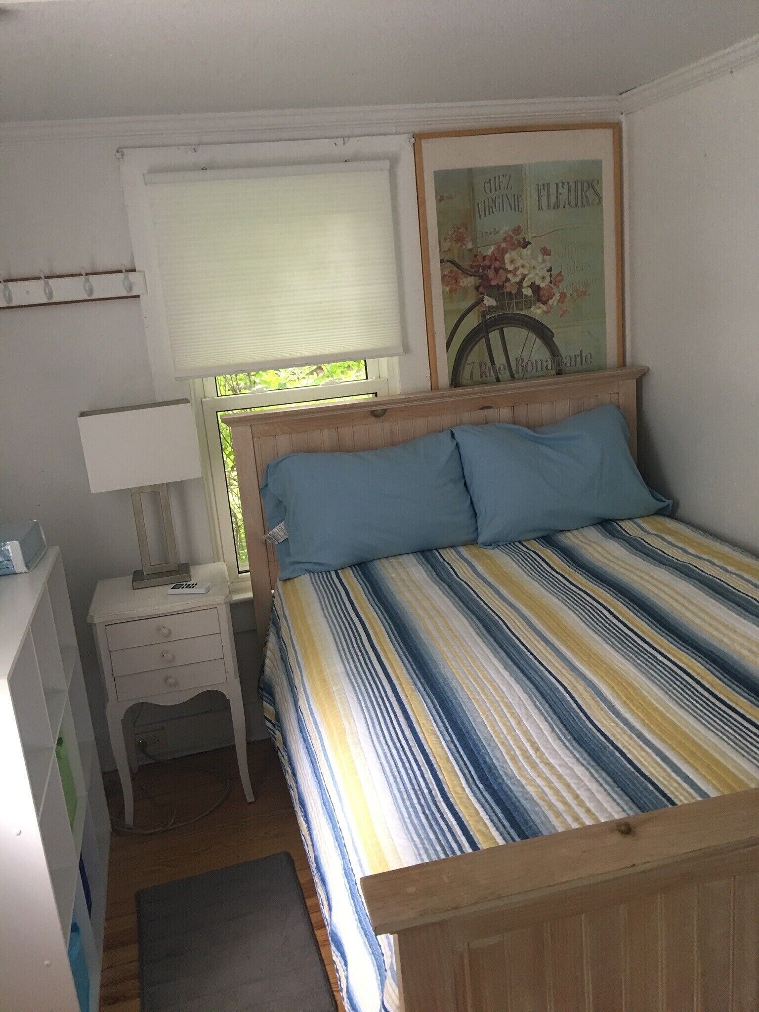 A small, neatly arranged bedroom with a single bed, a bedside table with a lamp, a framed picture on the wall, and a window with blinds.