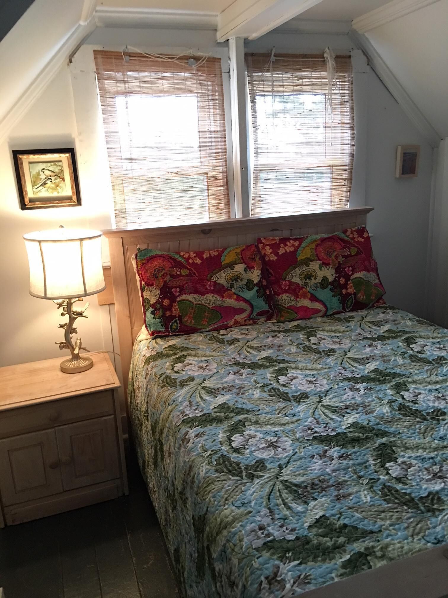 Quarto com cama, mesa de cabeceira, abajur e duas janelas com persianas.