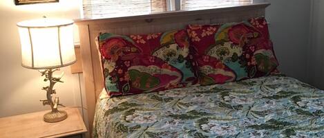 Quarto com cama, mesa de cabeceira, abajur e duas janelas com persianas.