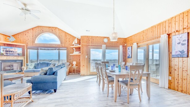 A spacious living area with a large sofa, dining table, and chairs, wooden walls, and large windows offering a view. - Oceanfront Dog Friendly Home in Salvo w/ Private Pool, Hot Tub, and Game Room (Salvo)