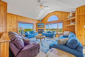 A spacious living room with a fireplace, large windows, and a ceiling fan. - Oceanfront Dog Friendly Home in Salvo w/ Private Pool, Hot Tub, and Game Room (Salvo)