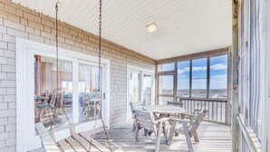 A covered outdoor area with a swing set, a wooden table with chairs, and large windows offering a view of the landscape. - Oceanfront Dog Friendly Home in Salvo w/ Private Pool, Hot Tub, and Game Room (Salvo)