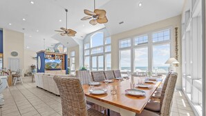 A spacious dining area with a long wooden table, wicker chairs, and large windows offering ocean views. - Premier Oceanfront Dog Friendly w/ Pool, Elevator, Hot Tub, Theater & Game Rooms (Hatteras)