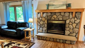 A living room with a stone fireplace, a black leather sofa, a wooden side table, and a rug with paw prints. - Cabin w/ gas fireplace, playground& AuTrain Lk Access (AuTrain)