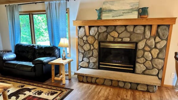 A living room with a stone fireplace, a black leather sofa, a wooden side table, and a rug with paw prints.