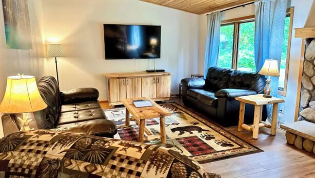 A living room with a sofa, a coffee table, a TV, and a stone wall. - Cabin w/ gas fireplace, playground& AuTrain Lk Access (AuTrain)