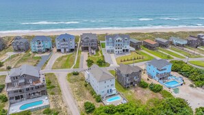 A coastal residential area with multiple two-story houses, a swimming pool, and a beach in the background. - Semi-Oceanfront Dog Friendly Home w/ Pool, Hot Tub, Renovated Kitchen, Game Room (Hatteras)