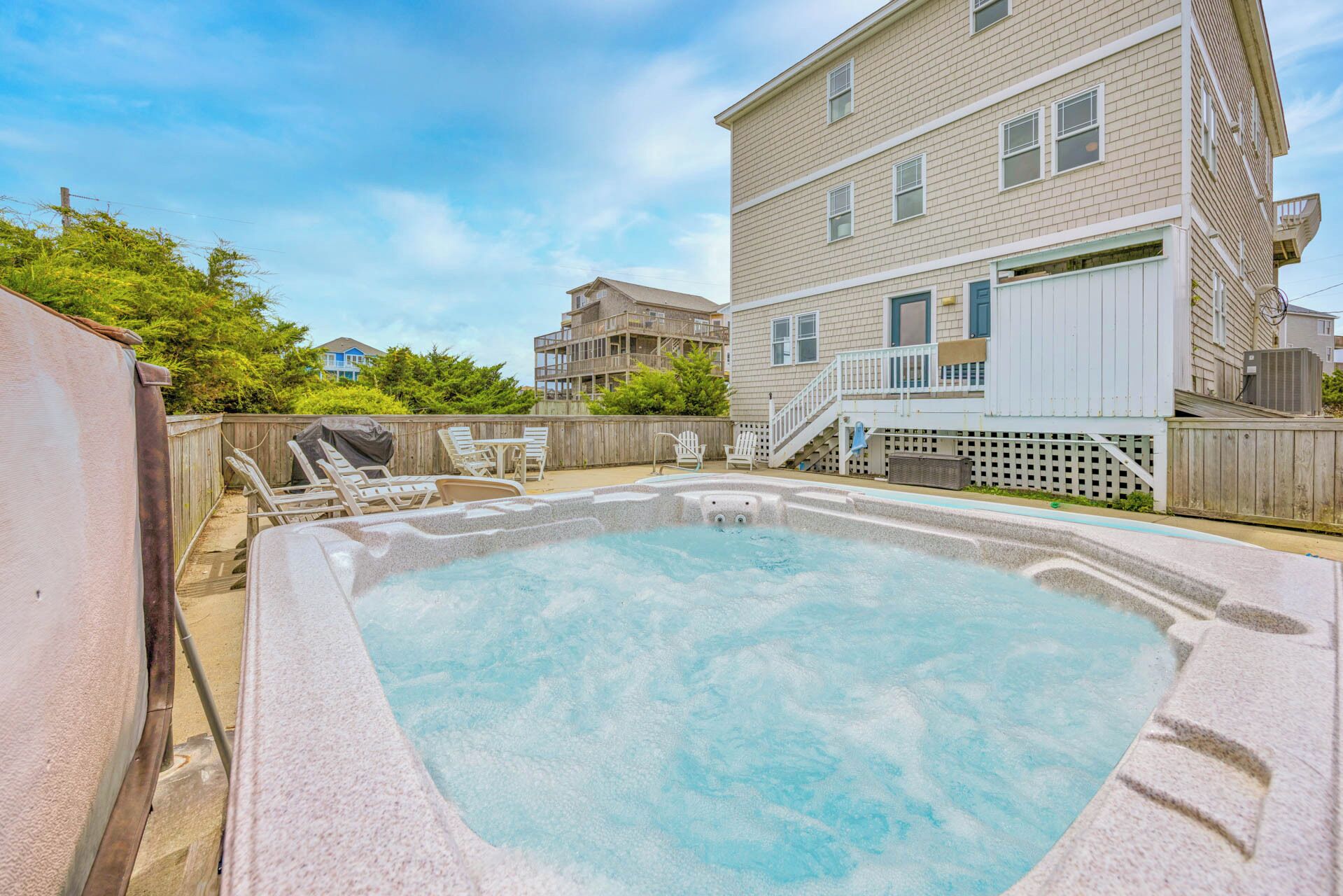 A hot tub in a backyard with a wooden deck, a covered patio, and a two-story building in the background.