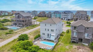 A residential area with multiple two-story houses, a swimming pool, and a beach in the background. - Semi-Oceanfront Dog Friendly Home w/ Pool, Hot Tub, Renovated Kitchen, Game Room (Hatteras)