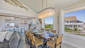 A modern kitchen with white cabinets, a dining area with wicker chairs, and a large window offering a view of a house. - Oceanfront Dog Friendly Home with Private Pool, Hot Tub, Direct Beach Access (Avon)
