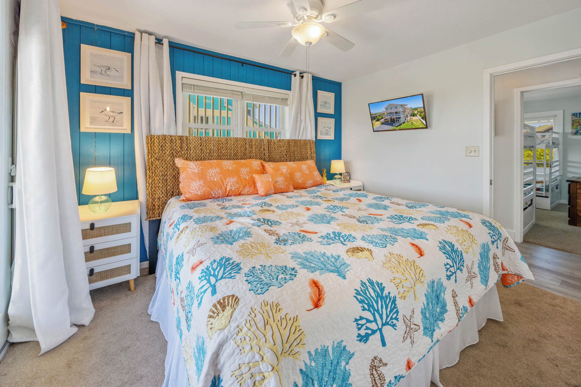 A bedroom with a bed, a ceiling fan, a window with curtains, a nightstand with a lamp, and framed pictures on the wall.