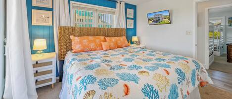 A bedroom with a bed, a ceiling fan, a window with curtains, a nightstand with a lamp, and framed pictures on the wall.