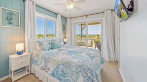 A bedroom with a bed, a nightstand, a ceiling fan, and a view of the beach through the window. - Oceanfront Dog Friendly Home with Private Pool, Hot Tub, Direct Beach Access (Avon)