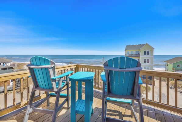 A wooden deck with two turquoise Adirondack chairs and a small table, overlooking a beach with a house in the background. - Semi-Oceanfront Dog Friendly Home w/ Private Pool, Hot Tub, Elevator, Game Room (Rodanthe)