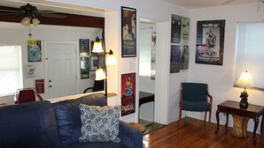 A living room with a dark blue sofa, a patterned throw pillow, a wooden coffee table, a chair, and a lamp. Posters adorn the walls, and a ceiling fan is visible.