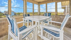 A covered outdoor patio with a white table and chairs, large windows offering a view of the beach and houses. - Semi-Oceanfront Home w/ Hot Tub, Smart TVs, Community Tennis/Pickleball Courts (Rodanthe)