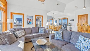 A spacious living room with grey sofas, a coffee table, and a dining area with wooden furniture. - Semi-Oceanfront Home w/ Hot Tub, Smart TVs, Community Tennis/Pickleball Courts (Rodanthe)