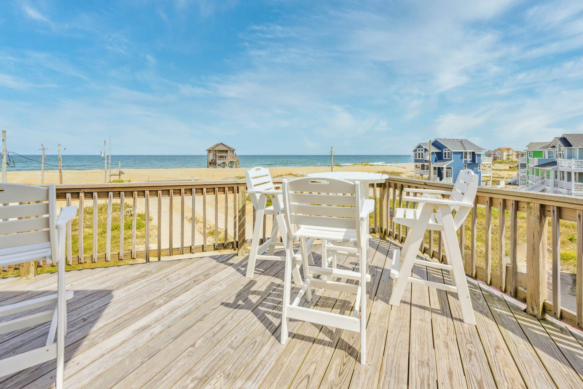 A wooden deck with white chairs and a table overlooking a beach with a lifeguard tower and houses in the background.