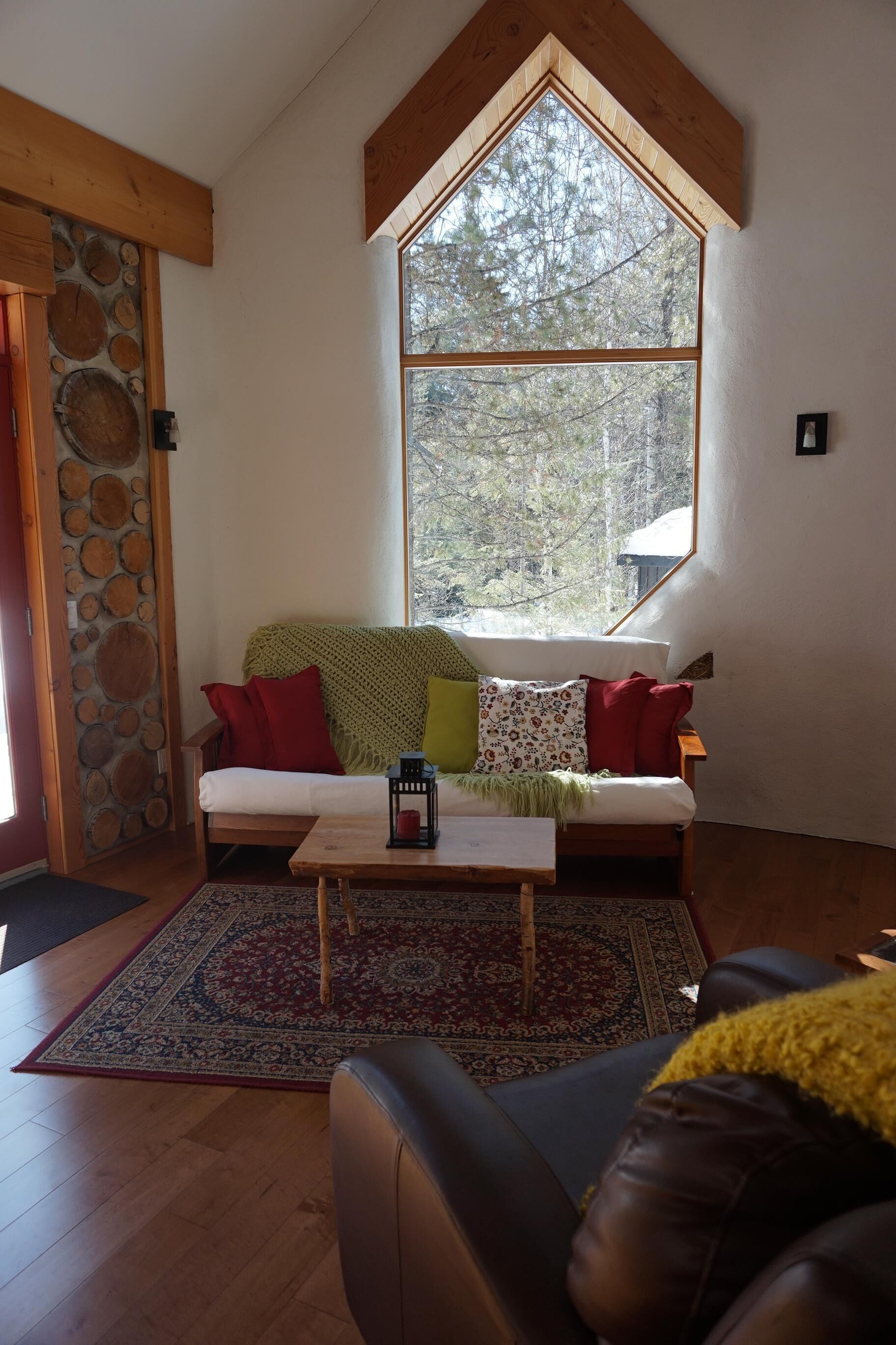 A cozy living room with a large window, a stone wall, a wooden ceiling, and a sofa with cushions.