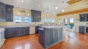 A spacious kitchen with dark wooden cabinets, a central island, and large windows providing views of the outdoors. - Oceanview Dog Friendly Home with Private Pool, Hot Tub, Elevator, Game Room (Avon)