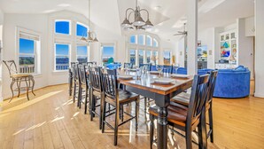 A spacious dining area with a long wooden table, multiple chairs, and pendant lighting. Large windows offer views of the cityscape. - Premier Oceanview Dog Friendly w/ Pool, Hot Tub, Elevator, EV Charger, Much More (Rodanthe)
