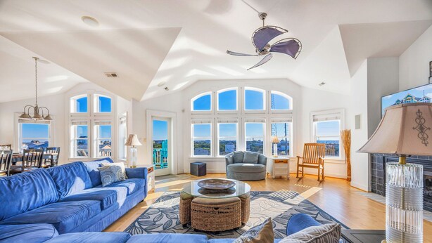A spacious living room with a blue sofa, a round coffee table, and a fireplace. Large windows offer a view of the cityscape. - Premier Oceanview Dog Friendly w/ Pool, Hot Tub, Elevator, EV Charger, Much More (Rodanthe)