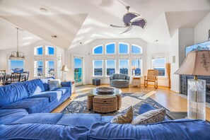 A spacious living room with a blue sofa, a round coffee table, and a fireplace. Large windows offer a view of the cityscape. - Premier Oceanview Dog Friendly w/ Pool, Hot Tub, Elevator, EV Charger, Much More (Rodanthe)