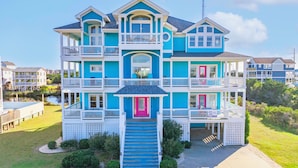 A multi-story beachfront house with balconies, a prominent pink door, and a clear view of the surrounding area. - Premier Oceanview Dog Friendly w/ Pool, Hot Tub, Elevator, EV Charger, Much More (Rodanthe)