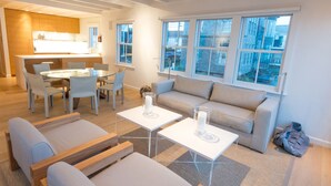 A modern living room with a sofa, two white coffee tables, and a dining area with a glass table and chairs. - Spectacular Beachfront West End Home  (Provincetown)