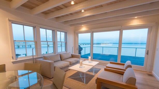 A spacious living area with a large window overlooking a body of water and a bridge. There is a sofa, a coffee table, and a dining table with chairs. - Spectacular Beachfront West End Home  (Provincetown)