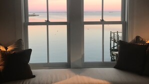 A room with a large window overlooking a calm body of water during sunset. There is a bed with pillows and a small table with chairs. - Spectacular Beachfront West End Home  (Provincetown)