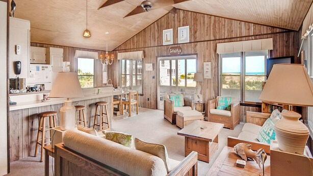 A spacious living area with a wooden ceiling, a large window overlooking a beach, a kitchen with white cabinets, and a seating area with a sofa and chairs.